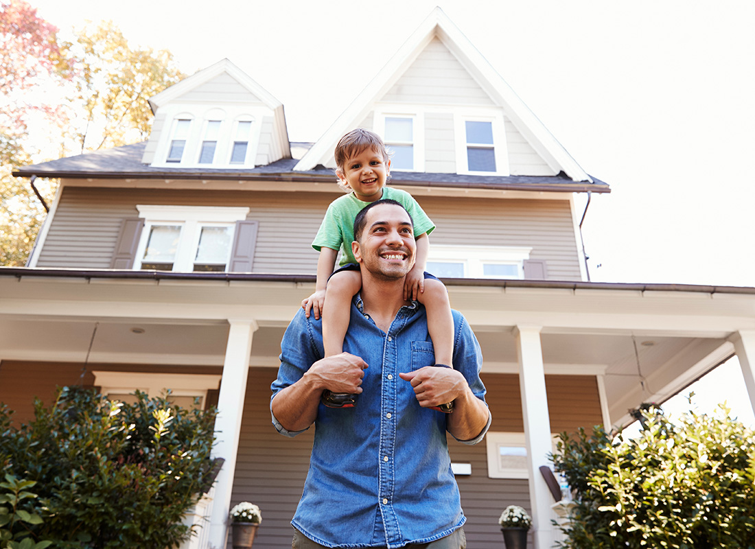 Personal Insurance - Portrait of a Cheerful Young Father Giving his Son a Piggyback Ride as They Stand Outside Their Two Story Home on a Sunny Day